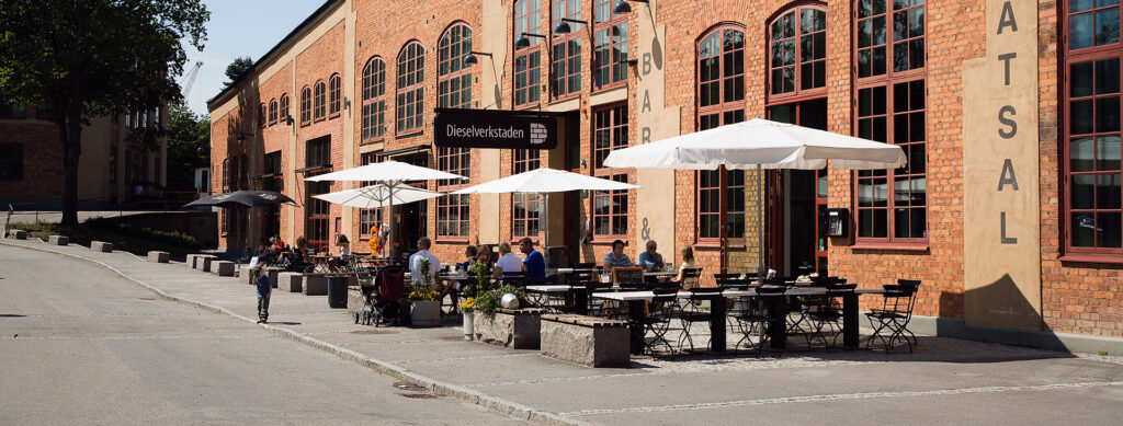 En tegelbyggnad som det är en servering framför med bord, stolar och parasoller. Det sitter några människor vid ett par av borden.