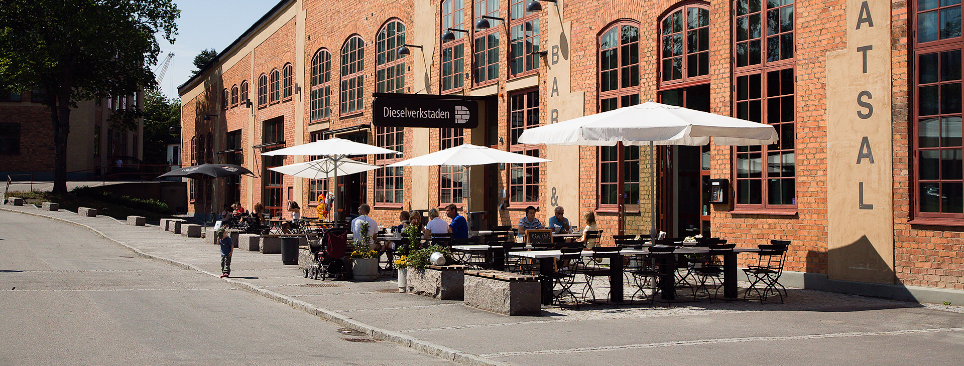 En tegelbyggnad som det är en servering framför med bord, stolar och parasoller. Det sitter några människor vid ett par av borden.