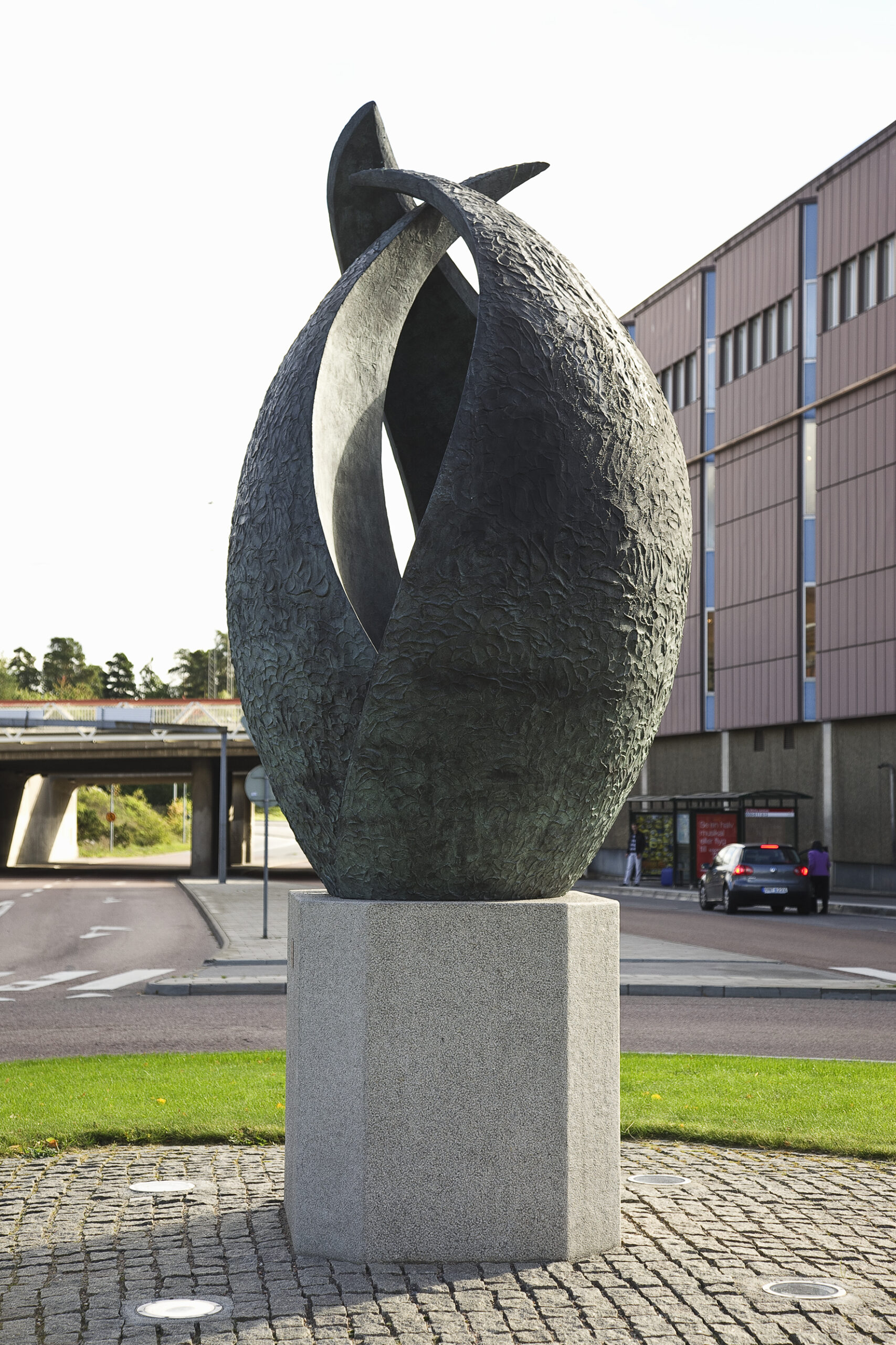 Grön bronsskulptur på grå betongsockel. Skulpturen liknar en jättelik blomknopp som är på väg att spricka upp. Cirka tre meter hög.