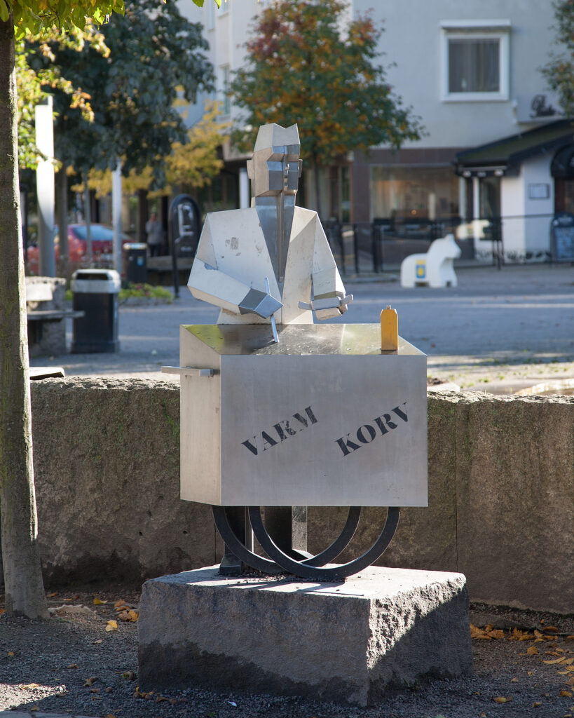 En skulptur av plåt av en korvgubbe med låda på magen. Hus och träd bakom.