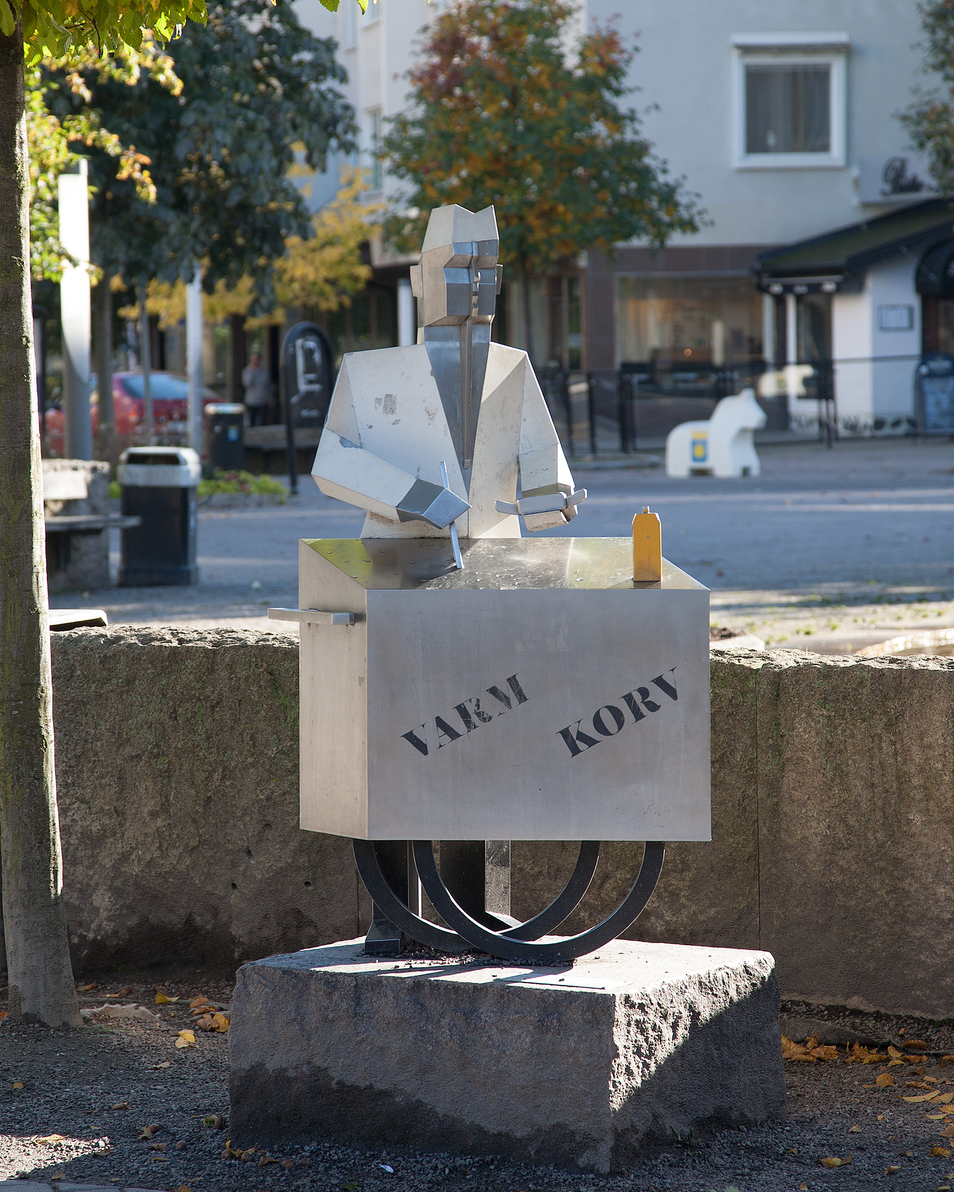 En skulptur av plåt av en korvgubbe med låda på magen. Hus och träd bakom.