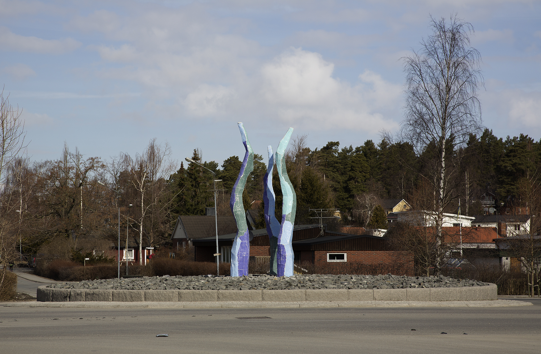 Tre höga pelarna med rundade former som klätts med mosaik i färgerna blått och grönt. Placerade i en rondell med sten.