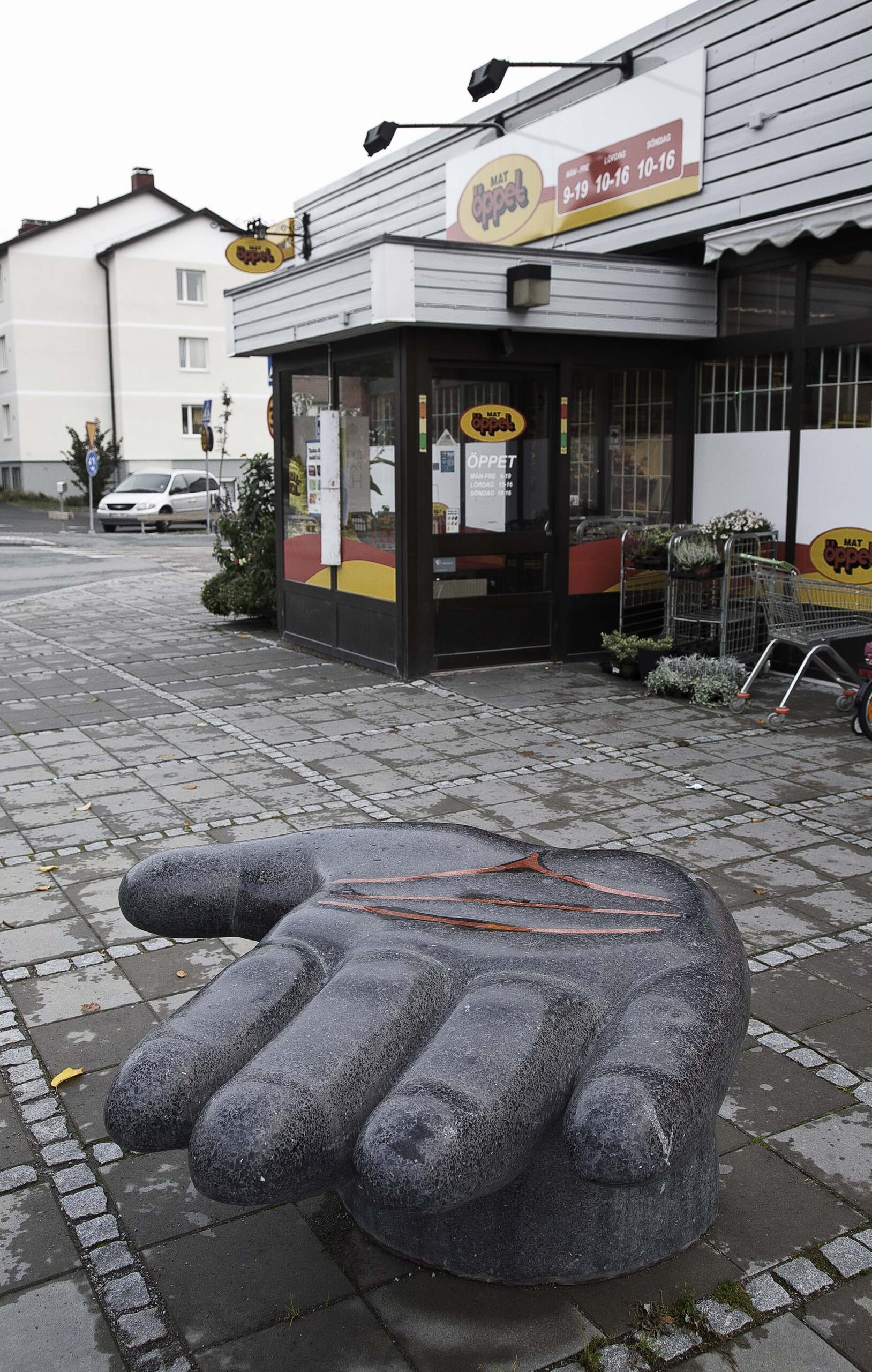 En skulptur av en öppen handflata av svart sten med röda linjer. Skulpturen är på stenläggning, en affär bakom.