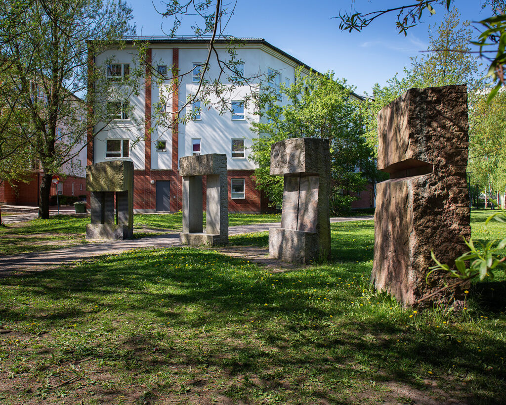 Fyra stora, rektangulära stående stenskulpturer som står på grönt gräs. Nyutspruckna träd och hus bakom.