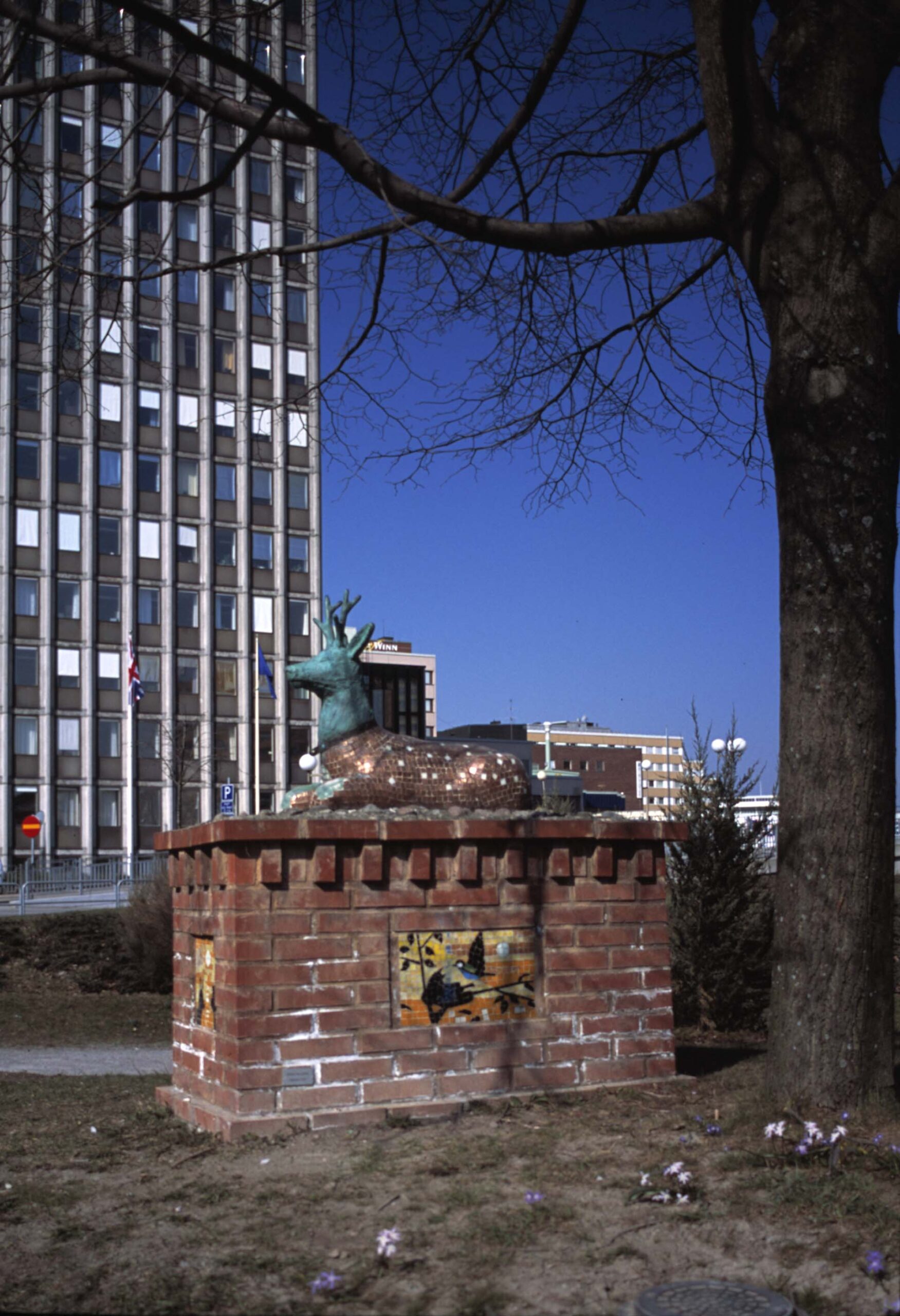 En skulptur av hjort i glittrig mosaik ligger på en fyrkantig sten som är omgiven av träd, gräs och hus.