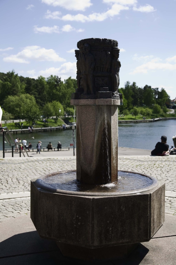 Ovanpå en pelare står figurer som liknar människor. Nedanför rinner vatten i ett stenkar. Människor och vatten bakom.