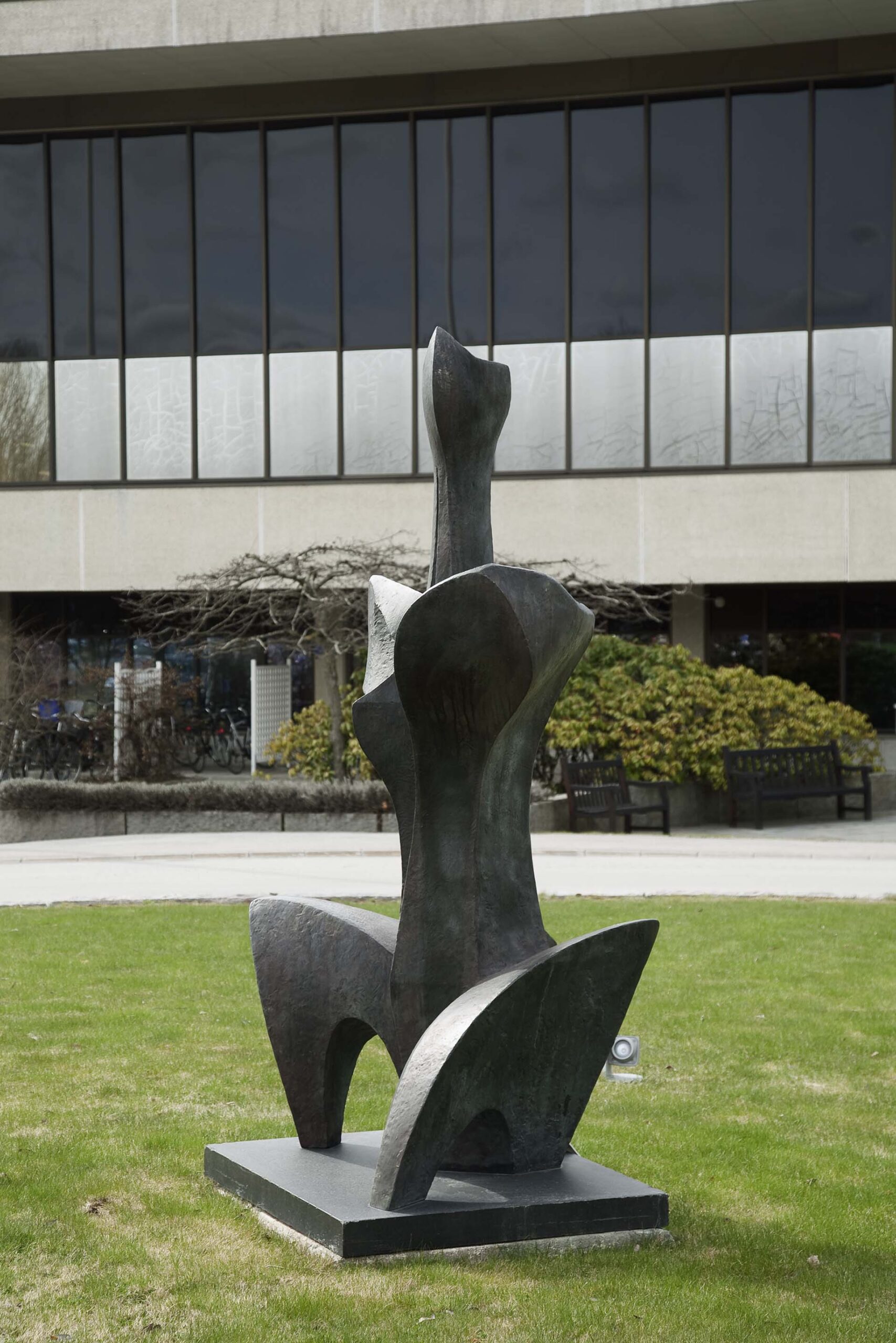 Abstrakt hög skulptur i brons som liknar en sittande person på en gräsmatta. Byggnad bakom.