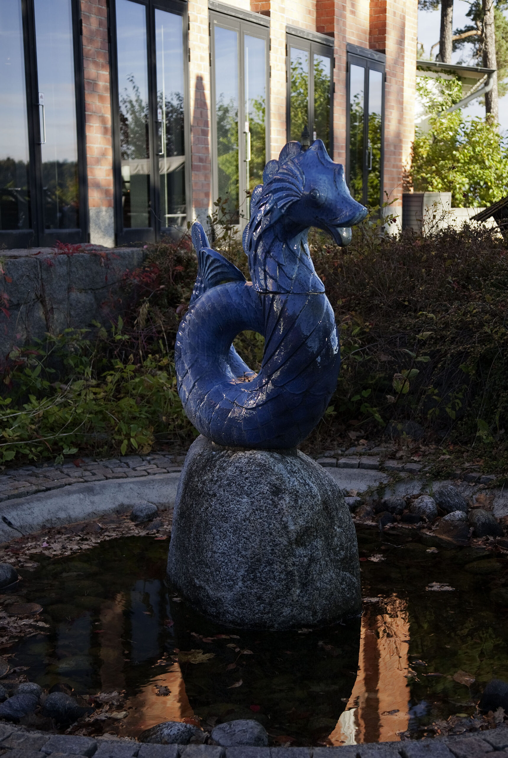En skulptur av en blå sjöhäst på en en sten. Växtlighet och vatten bakom.