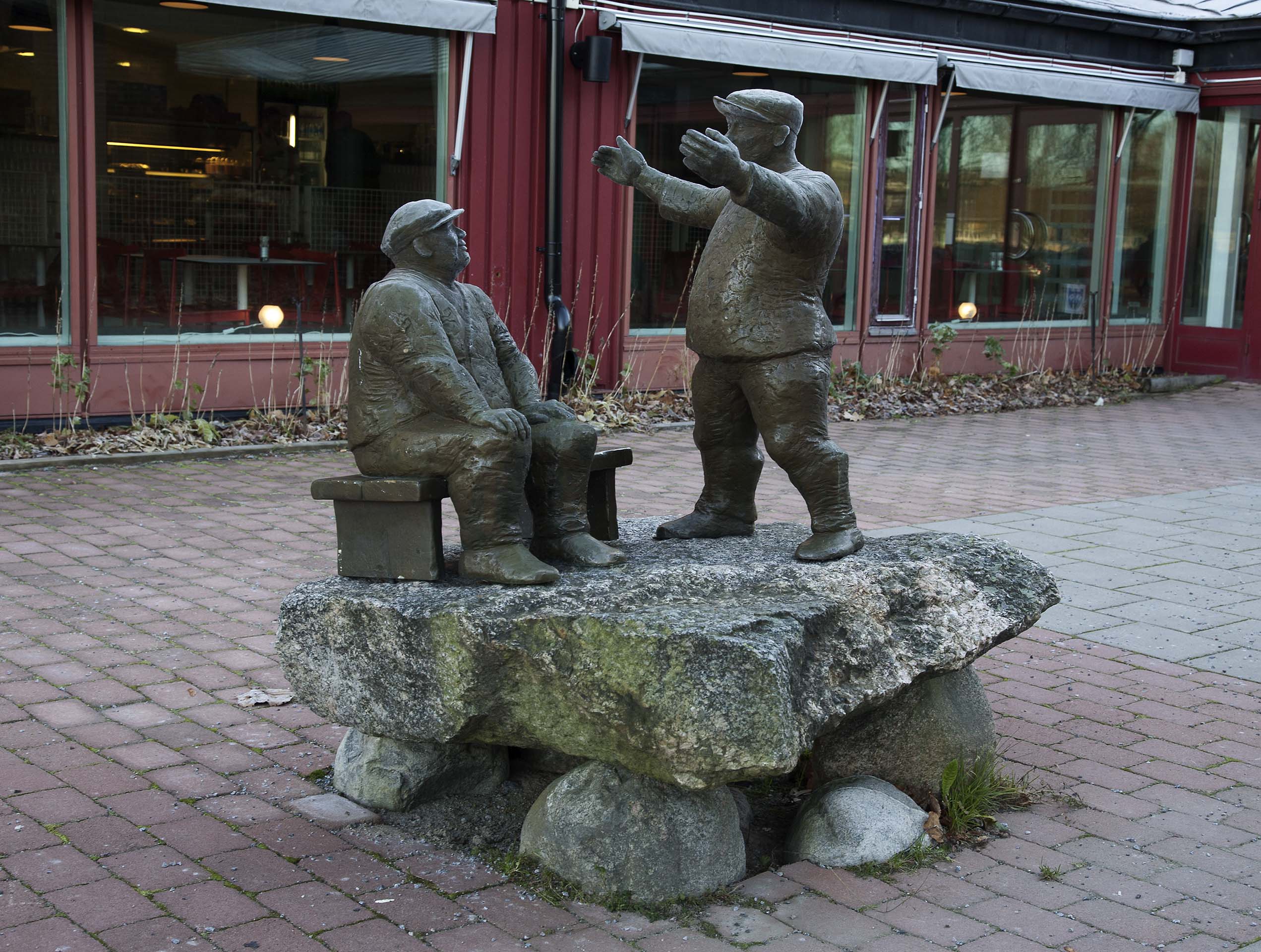 Två män i ett samtal. Den ena sitter på en bänk och den andra står upp och måttar upp ett avstånd mellan händerna.