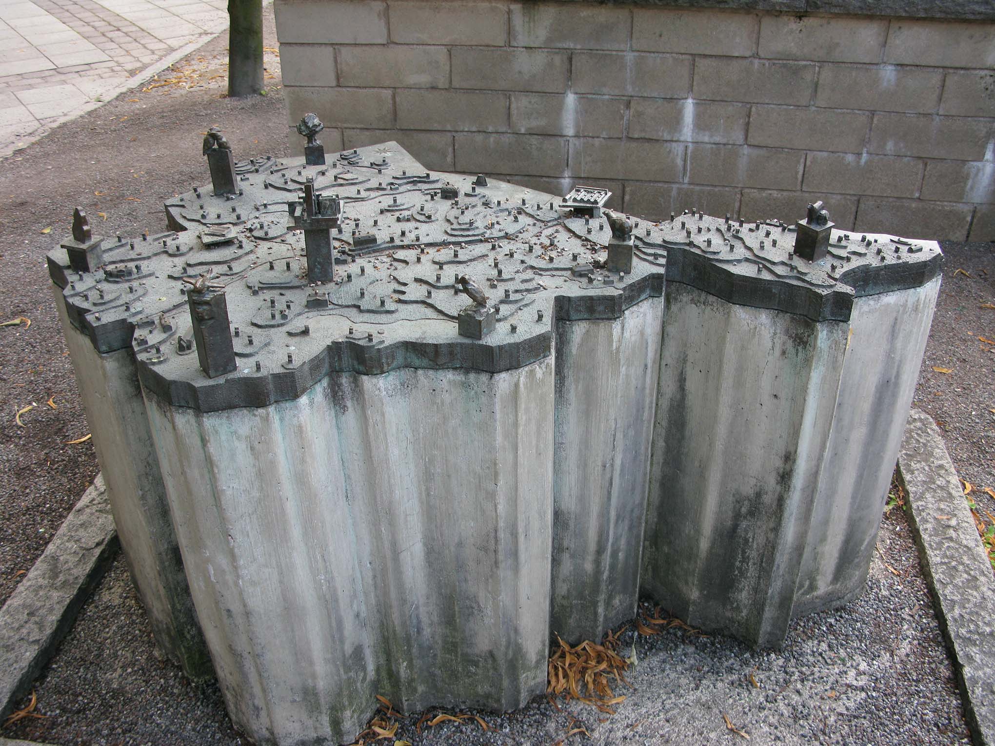 En modell i brons med figurer och hus på en sockel utomhus.