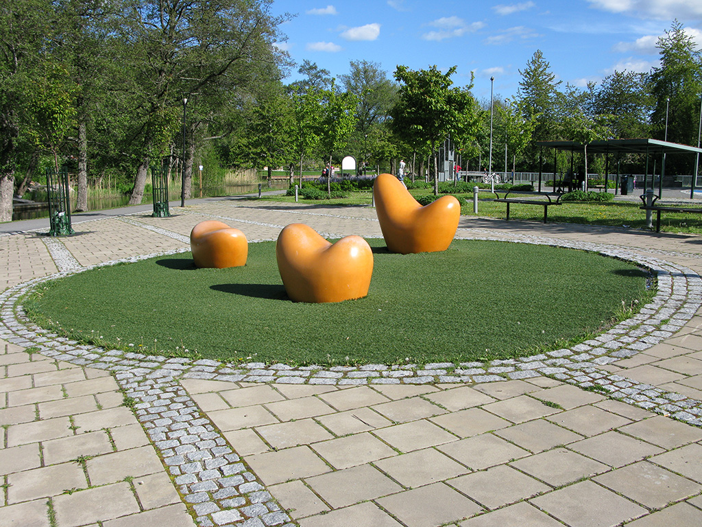 Tre brandgula figurer med mjuka former, alla är olika på en rundel av konstgräs.