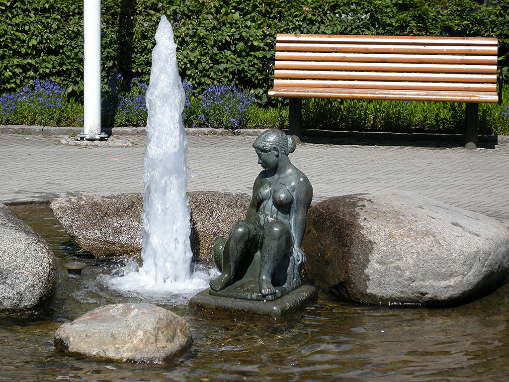 En skulptur av en naken flicka som sitter i en fontän med porlande vatten stenar.