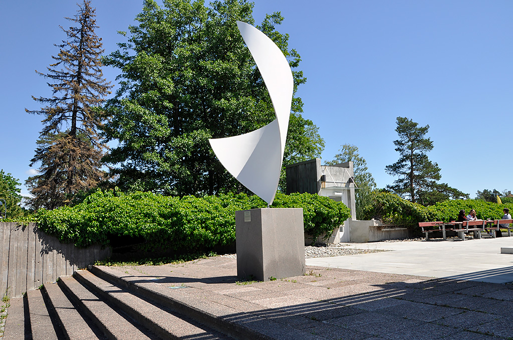 En skulptur av hög vit böjd form. En trappa och en uteplats med grönska bakom.