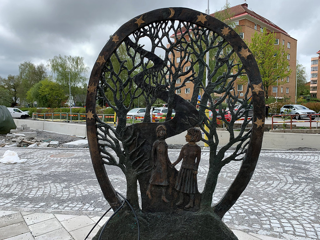 Två kvinnor håller varandra i händerna. De står i en cirkel, runtomkring utvecklar sig träd och grenar och bildar ett mönster, likt ett stort gallerverk. Stjärnor och månar runtom alltihop. Gatumiljö.