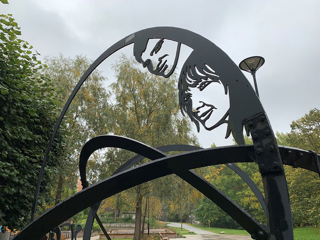 Skulptur i järn av ett ansikte av en man och en kvinna. Träd och buskar och himmel bakom.