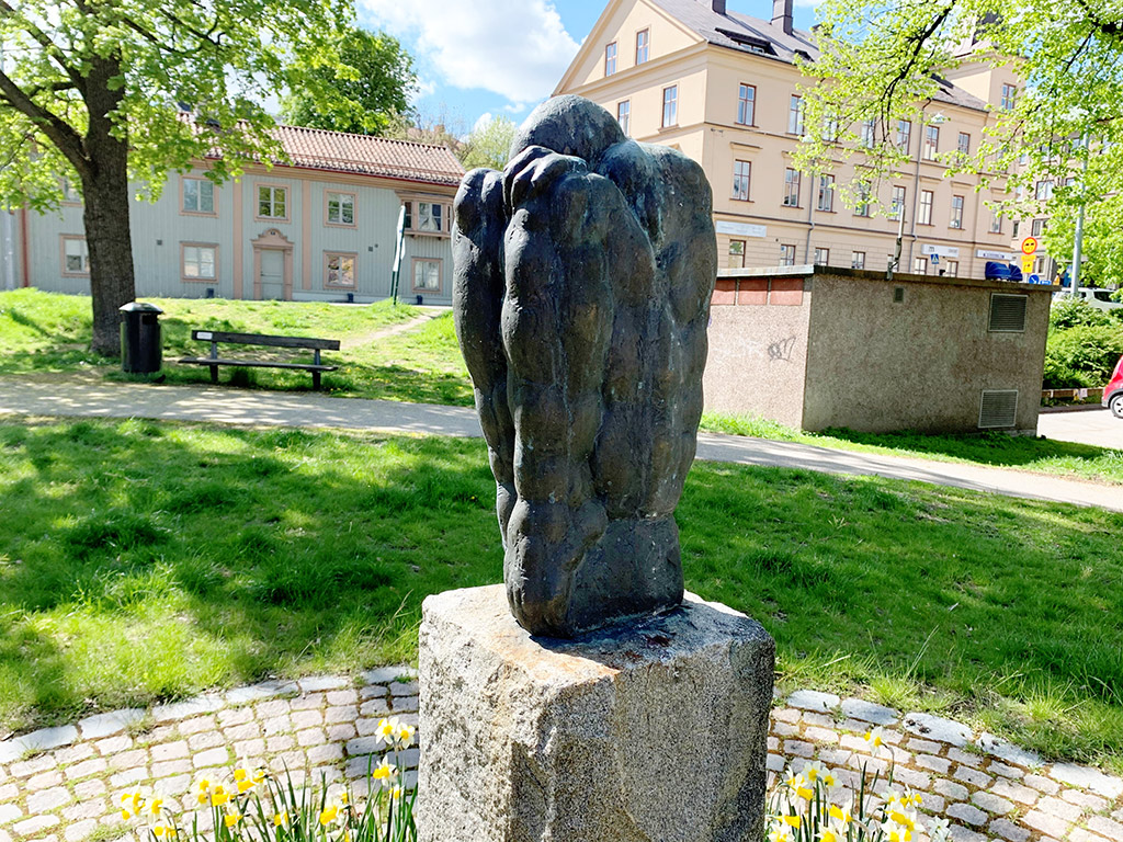 En skulptur av en man sitter med uppdragna knän och gömmer huvudet mellan benen. Hus och grönska bakom.