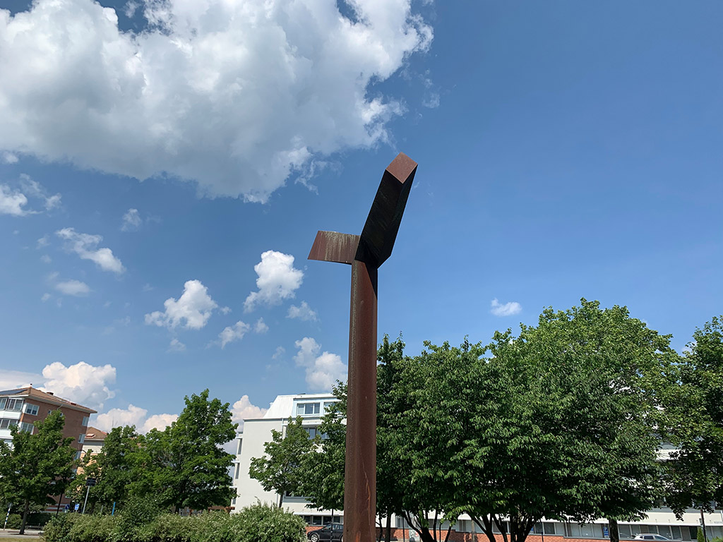 Skulptur av rostigt järn framför en vit byggnad och grönskande träd..