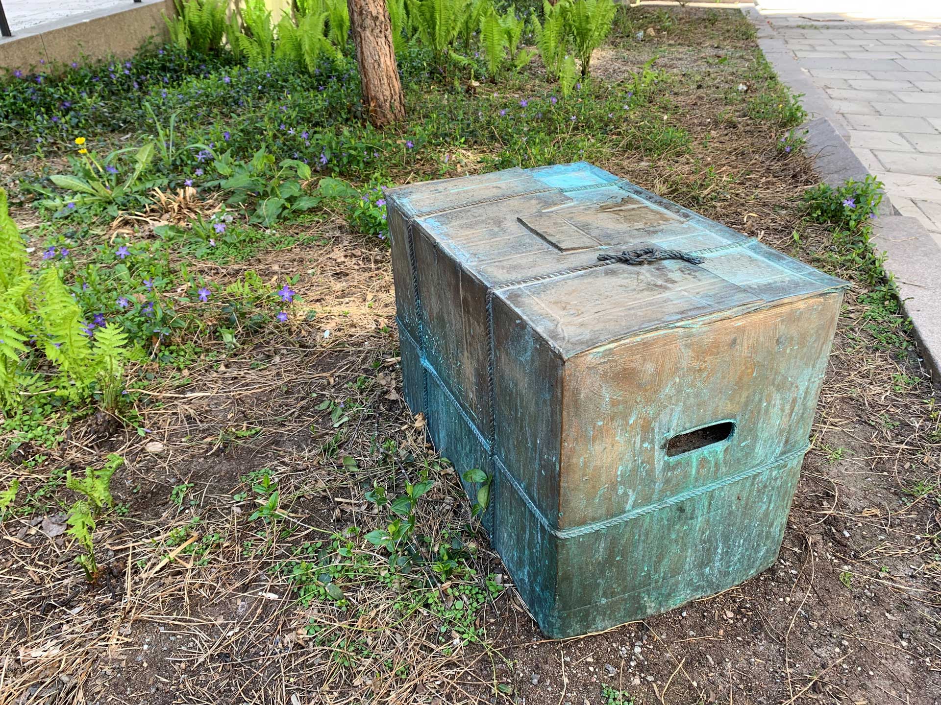 En fyrkantig låda av brons som står i en rabatt.