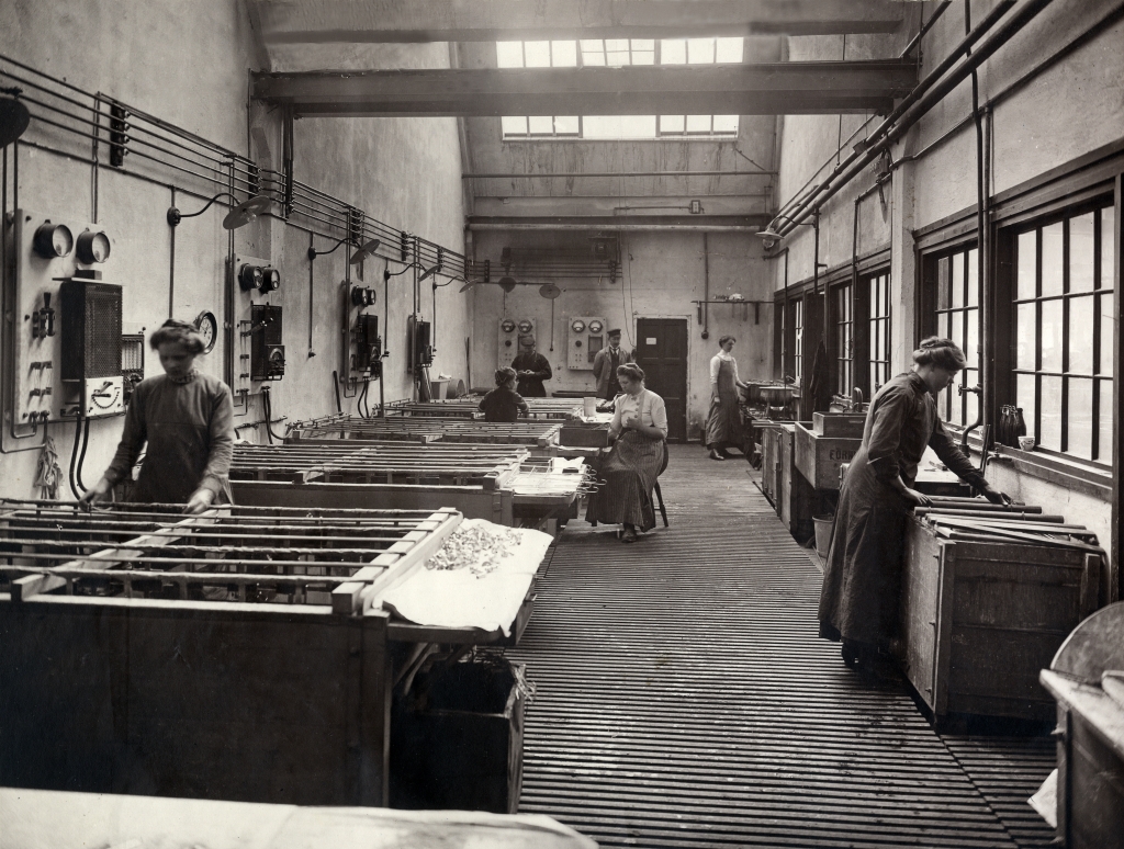 Svartvitt fotografi, några kvinnor och ett par män arbetar i en verkstad.