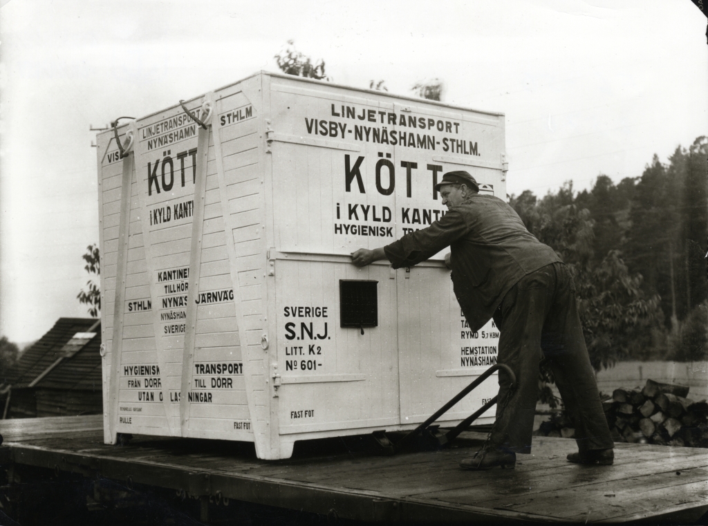 Svartvitt fotografi, man står på trädäck och flyttar en stor vit kontainer märkt med svart text.