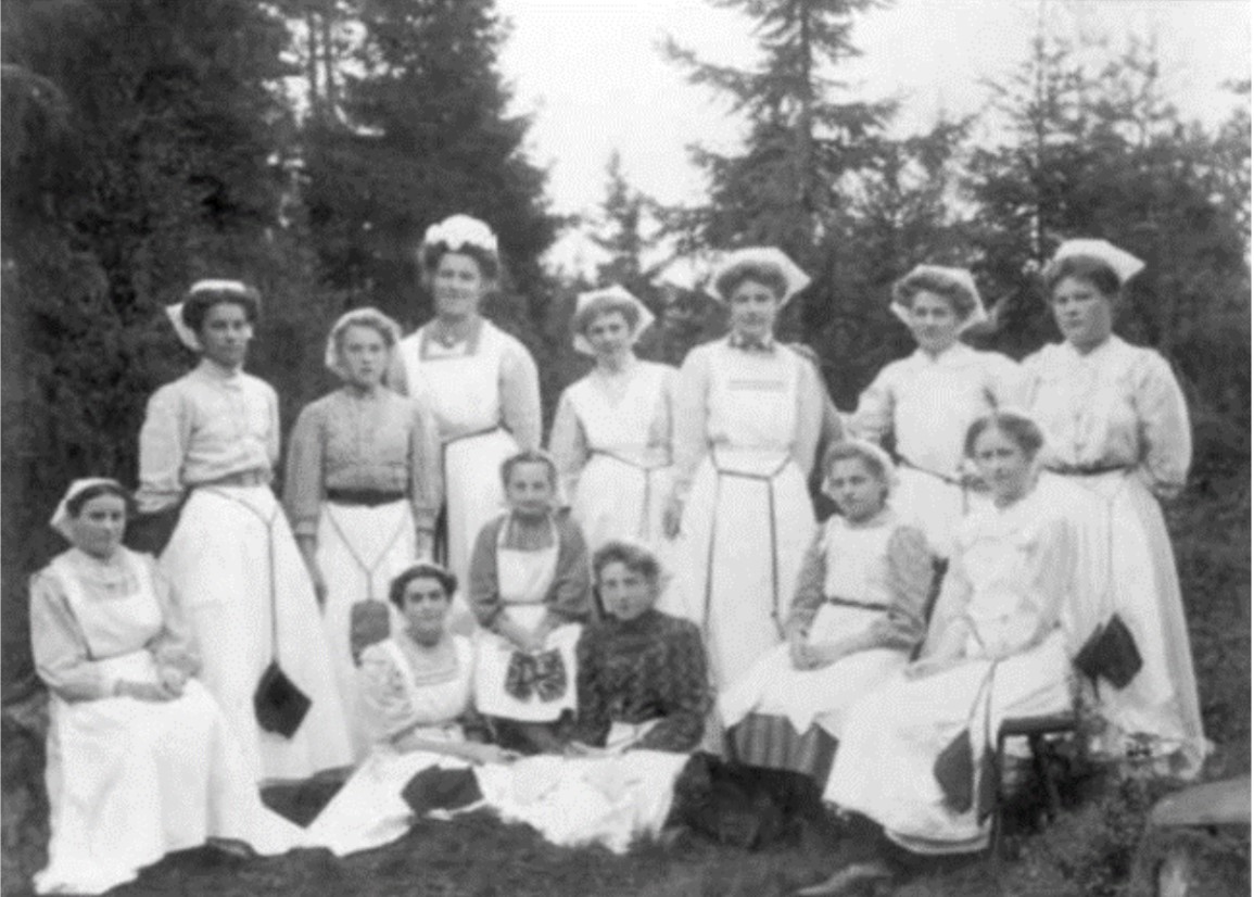 Svartvitt foto med tretton kvinnor som ställt upp sig för fotografering mot en fond av granar.