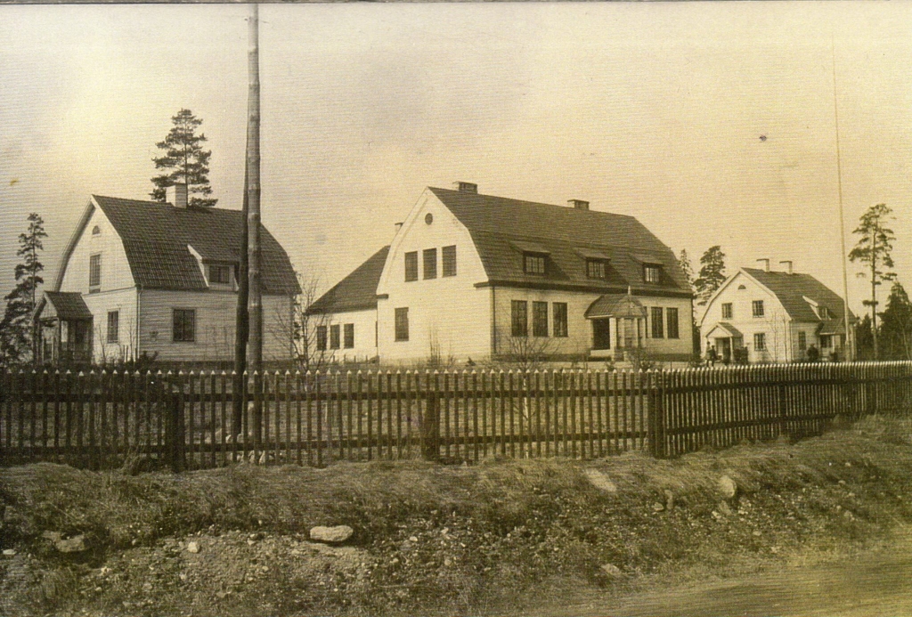 Svartvit bild med fyra vita byggnader. En stor i mitten omgiven av mindre hus.