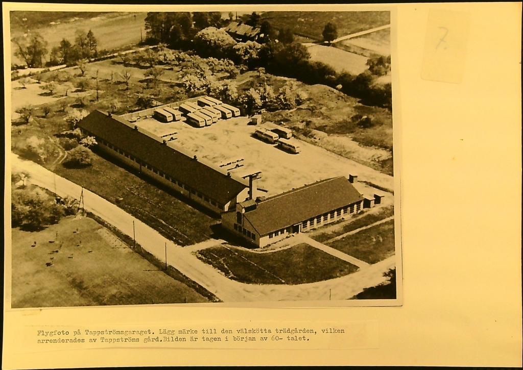 18 bussar står uppställda på en stor gård. Två långa garagebyggnader i vinkel ramar in gården. Längst bort i bilden syns träd i rader och annan växtlighet.