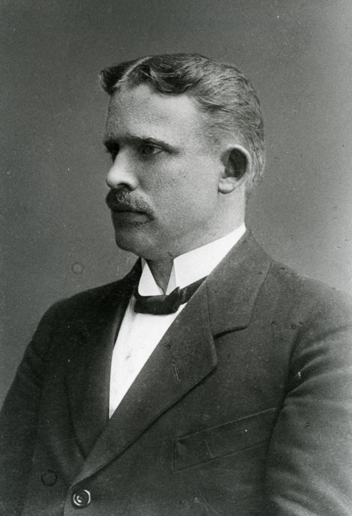 Svartvitt fotografi av en man i profil som bär fluga och mustasch.