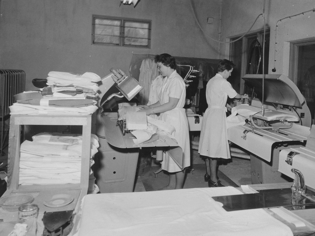 Svartvitt fotografi, två kvinnor stryker och hänger tvätt i tvätteriet.