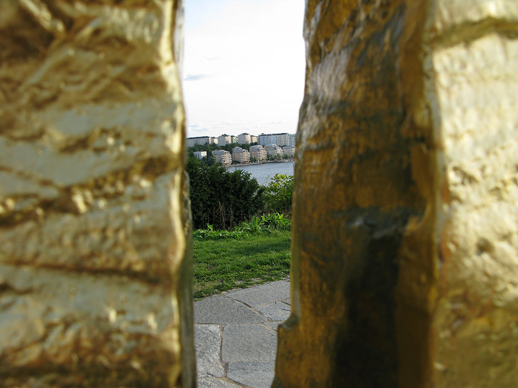 Utsikt över gräs, vatten och hus genom en öppning mellan två gyllene block.