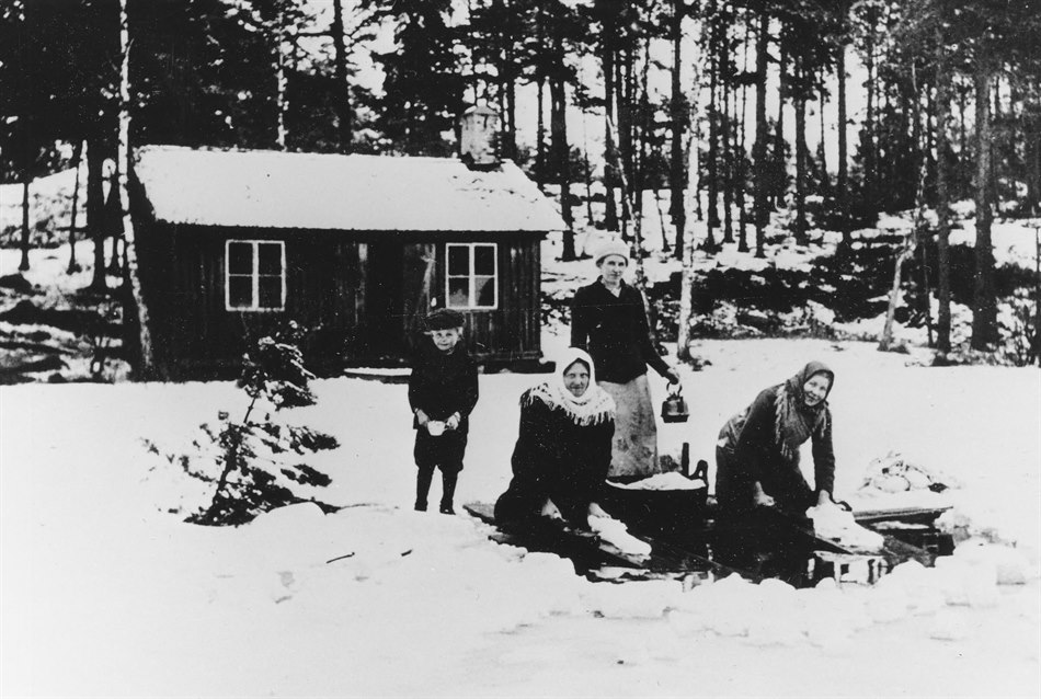Två kvinnor står på knä och tvättar i en vak i isen strax intill land. Ett barn står bredvid. En kvinna står vid en träbalja och håller en kanna i handen.