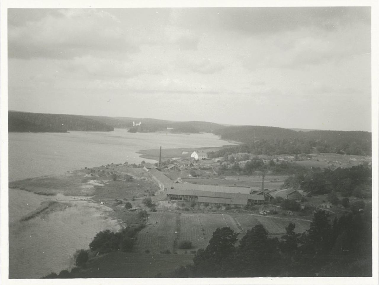 Vy från hög höjd över vatten till vänster och land till höger där några hus syns i mitten, bland dem långa lador och ett vitt hus på en höjd. I bakgrunden skymtar en vit byggnad längst in i viken.