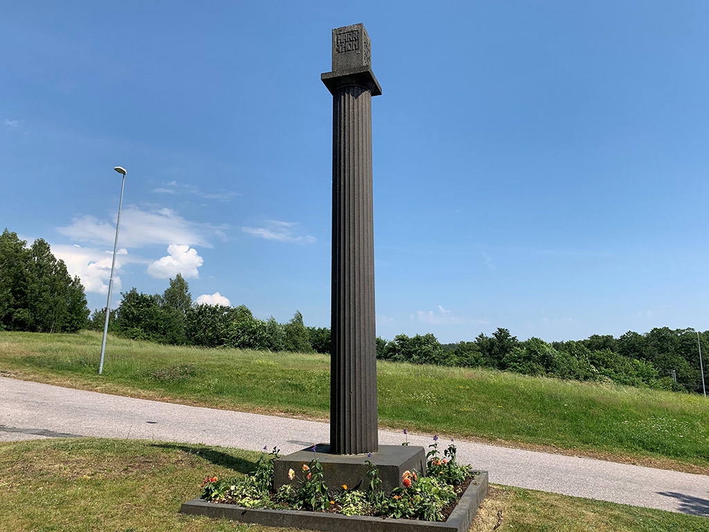 Hög räfflad stenpelare med blå himmel och grönska bakom.
