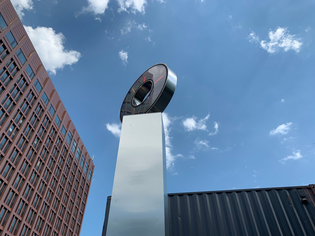 Rund klocka av rostfritt stål på en hög blänkande pelare. Blå himmel med molntussar och höga byggnader.