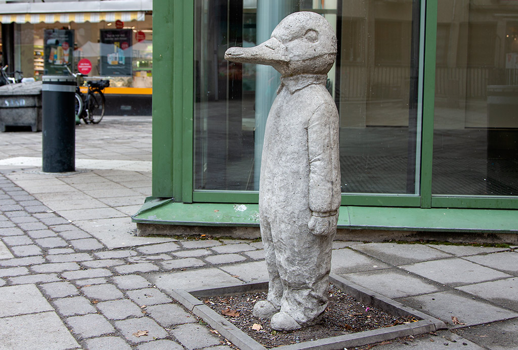Skulptur av en and i betong framför ett fönsterglas. Utomhus.