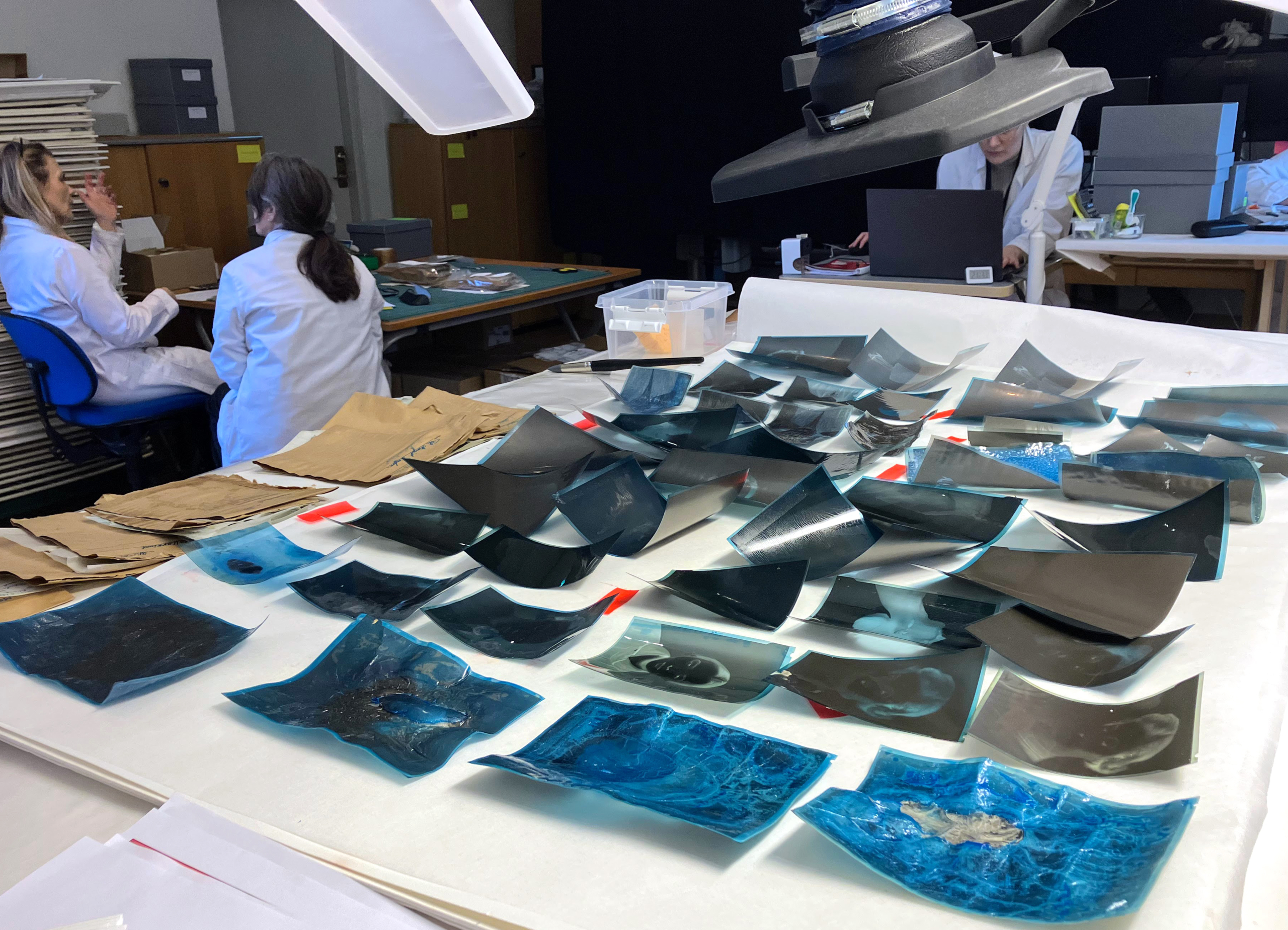 Negativ ligger utspridda på ett bord. Två personer sitter vid ett skrivbord i bakgrunden.