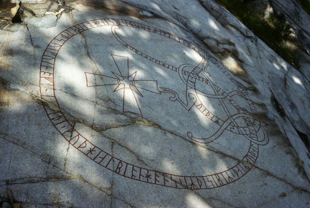 En berghäll med runor i en slinga. I mitten ett kors.