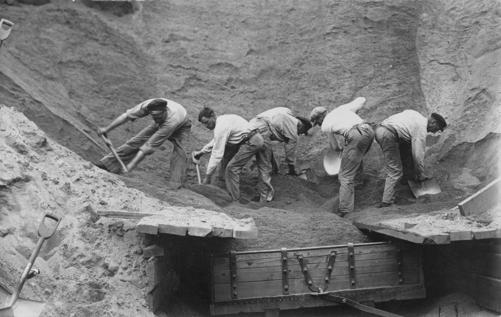 Fem män står böjda över en sandgrop och gräver upp sand.