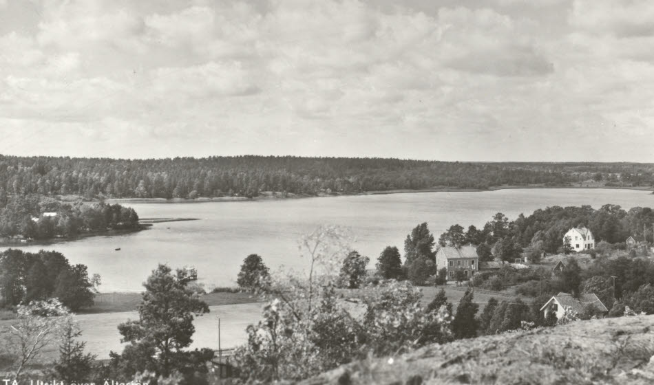 Svartvitt foto taget från en bergsknalle. Här syns idrottsplatsen, sjön, skog och tre hus.