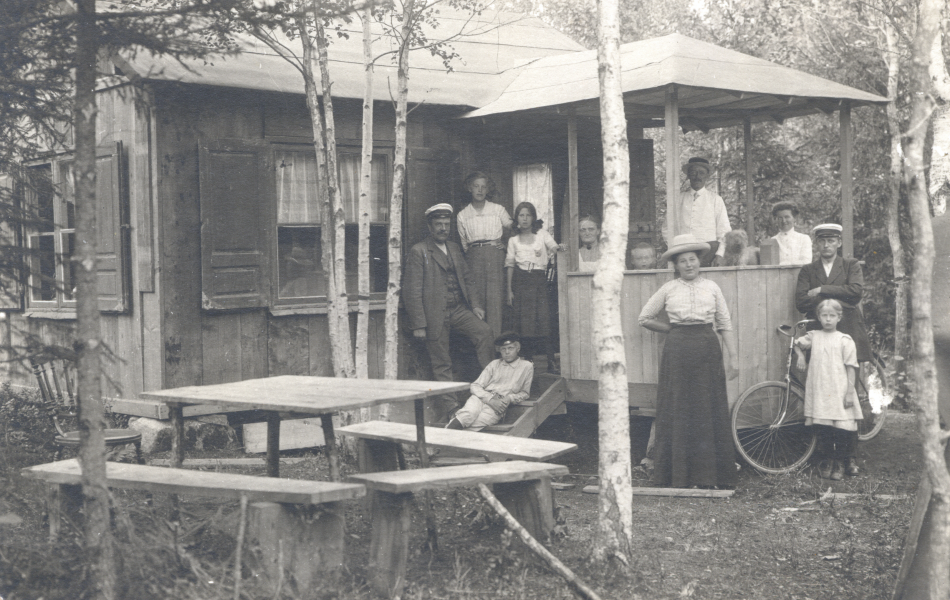 Svartvitt foto av 10 personer , en hund och en cykel som står uppställda på och runt verandan på ett hus.