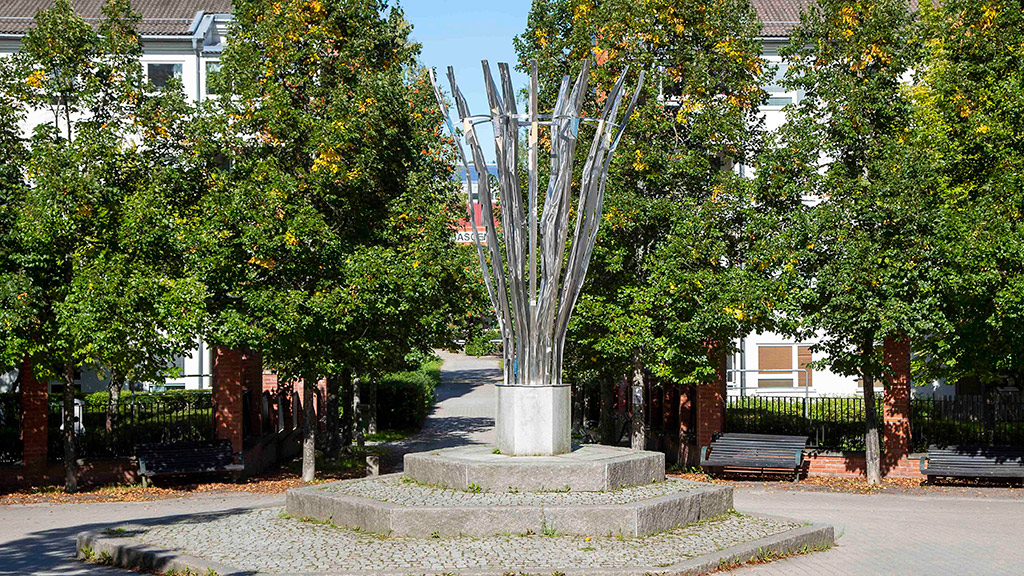 Glänsande och silverfärgade smala spröt som vidgar sig utåt upptill. Står på en sexkantig graderad plattform. Gröna träd bakom.