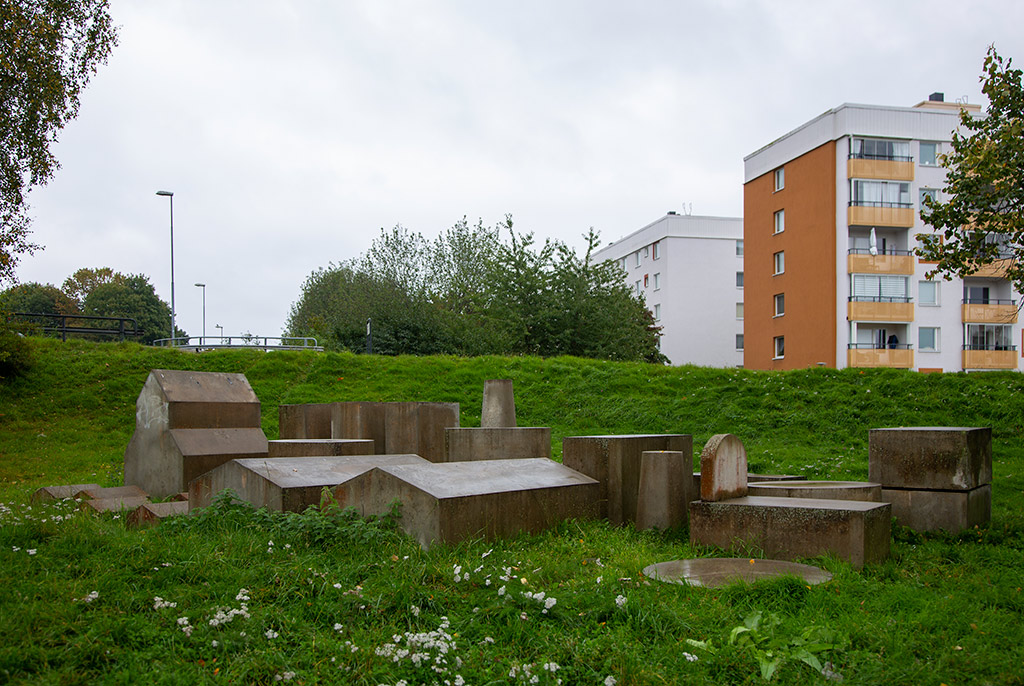 En samling med små gråa hus i en grässlänt. Bebyggelse med höghus bakom.