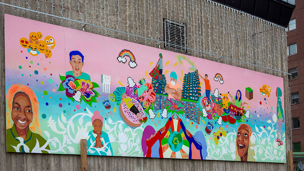 En stor avlång färgglad målning på en grå vägg. I målningen finns människor som skrattar, står och går. Regnbågar, smileys, höghus och växtlighet.