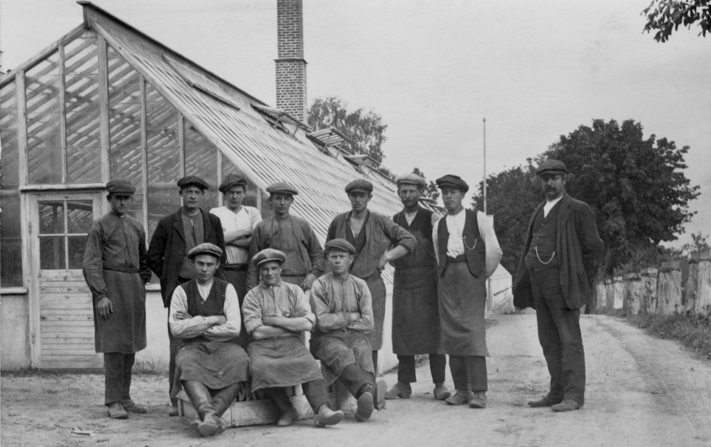 Svartvitt foto av 11 män med keps uppställda framför ett växthus med hög skorsten.