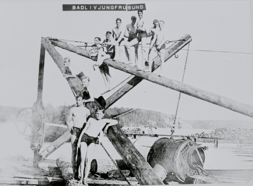 Svartvitt foto av elva pojkar i badbyxor som klättrar på en kran intill vattnet.