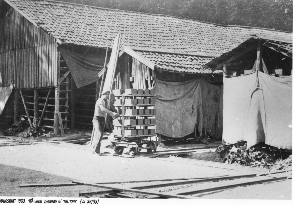 Svartvitt foto av en man i keps och kortärmad skjorta som skjuter en vagn med tegelstenar på en tågräls.