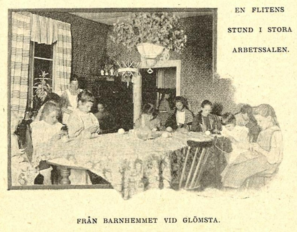 Ett tiotal flickor sitter runt ett stort bord i ett rum och handarbetar. Garnnystan på bordet. Texten lyder: En flitens stund i stora arbetssalen. Från barnhemmet vid Glömsta.