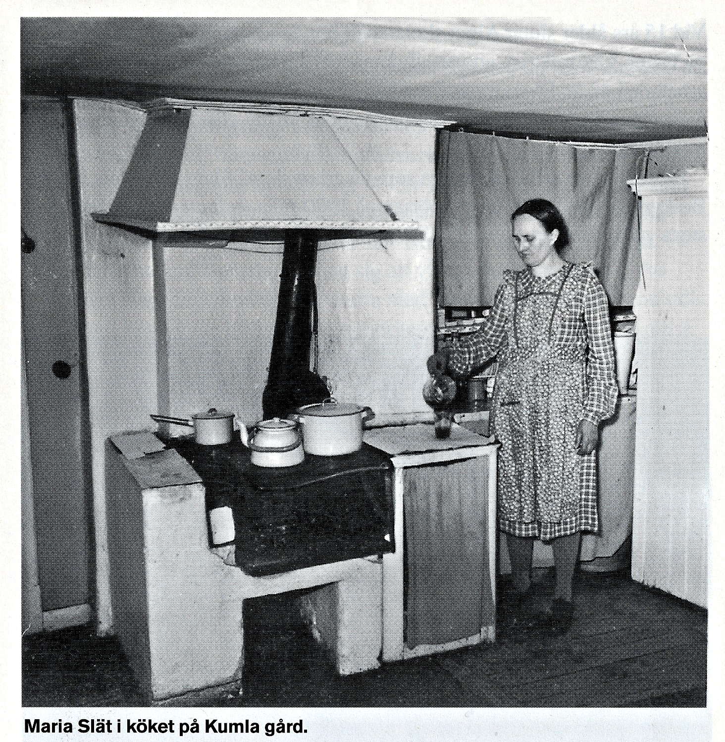 Svartvitt foto av Maria i det ålderdomliga köket med vedspis.