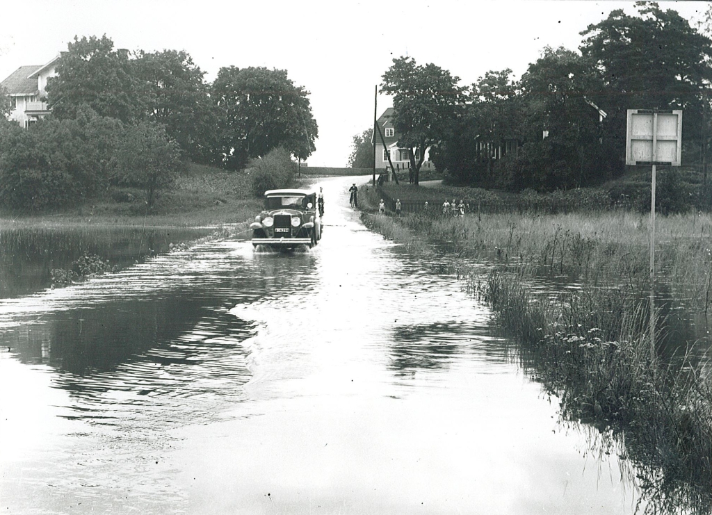 En ålderdomlig bil kör genom en helt översvämmad väg över en äng.