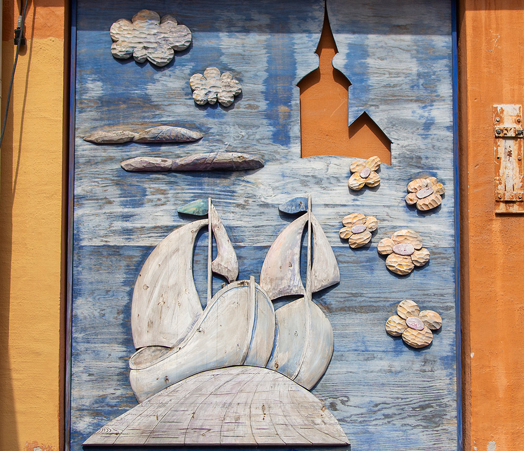 Bemålat konstverk av trä. Två vita segelbåtar syns på ett blått hav med gråa kobbar och skär och gula blommor. Längst upp till höger ligger en brun kyrka.